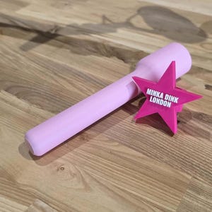 May include: A light pink cylindrical handle attached to a bright fuchsia star-shaped emblem. The star features the white text MINKA DINK LONDON printed in the center. The object has a smooth, matte finish and a uniform, solid color design.