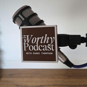 May include: A black RØDE microphone featuring a square brown flag attached to the side. The flag displays the white text "The Worthy Podcast WITH CHANI THOMPSON" in a stylized font. A black mounting bracket and a blue cable are also visible.