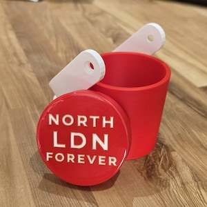 Puede incluir: Un recipiente cilíndrico rojo con una tapa roja a juego que presenta el texto en relieve blanco NORTH LDN FOREVER. Dos accesorios blancos planos en forma de asa con orificios circulares sobresalen de la parte posterior del vaso rojo.
