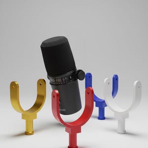 May include: A black Shure microphone positioned behind four U-shaped microphone mounts in gold, red, blue, and white. Each mount features a cylindrical base and two upward-extending arms with circular holes at the top for mounting hardware.