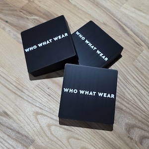 May include: Three black square boxes with the text "WHO WHAT WEAR" in white, arranged on a wooden surface. The boxes are evenly spaced and slightly angled, creating a visually appealing arrangement.