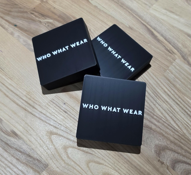 May include: Three black square boxes with the text "WHO WHAT WEAR" in white, arranged on a wooden surface. The boxes are evenly spaced and slightly angled, creating a visually appealing arrangement.