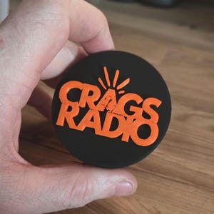May include: A black circular item with the words "CRAGS RADIO" in orange, with a stylized sunburst design above the word "RADIO". The item is held in a hand.