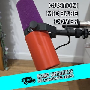Peut inclure: Un cache de base de microphone personnalisé avec un pare-vent en mousse violet et une base orange. Le texte "CUSTOM MIC BASE COVER" est affiché en blanc, en style pochoir. Le microphone est monté sur un bras noir.