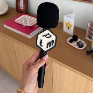 May include: A black handheld microphone featuring a large, black foam windscreen and a hexagonal white flag attached to the handle. The flag displays the black, pixelated letters 'p', 'l', and 'm' arranged in a vertical and horizontal layout.