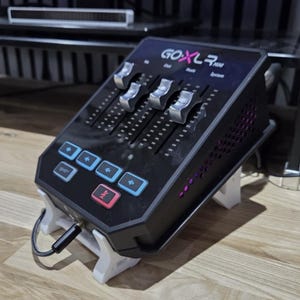 May include: Black audio mixer with silver sliders and blue and red buttons. The mixer has the text "GOXLR Mini" on the top. It is supported by a white stand and has a black cable plugged in.