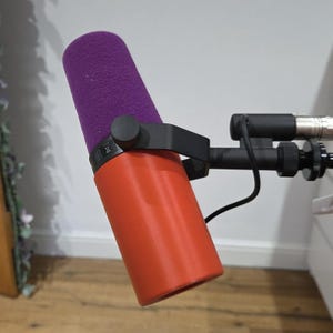 May include: A red and black microphone with a purple foam cover. The microphone is mounted on a black arm and connected to a silver cable. The microphone is angled slightly to the left.