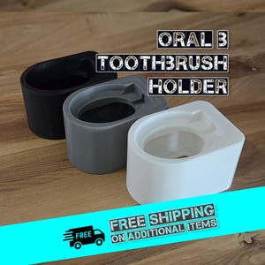 Könnte beinhalten: Drei Oral-B Zahnbürstenhalter in Schwarz, Grau und Weiß, auf einer Holzoberfläche angeordnet. Die Halter haben eine geschwungene Form. Der Text "ORAL B TOOTHBRUSH HOLDER" ist über den Haltern zu sehen.