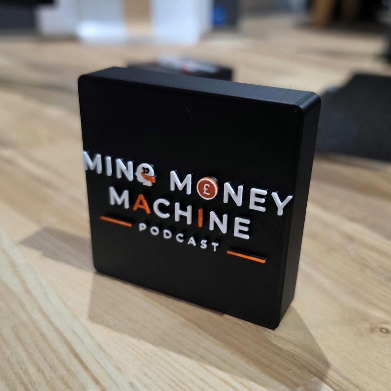 May include: A black rectangular block with the words "MING MONEY MACHINE PODCAST" in white and orange lettering. The design includes a pound symbol. The block is sitting on a wooden surface.
