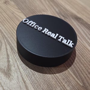May include: A black, circular object with the text "Office Real Talk" in white, raised lettering. The object is set against a wooden background.