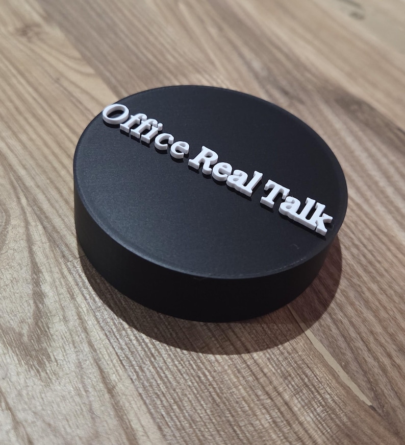 May include: A black, circular object with the text "Office Real Talk" in white, raised lettering. The object is set against a wooden background.