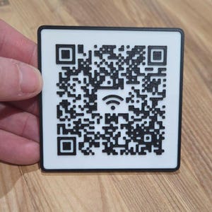 Op de afbeelding: Een vierkante QR-code in zwart en wit met een wifi-symbool in het midden. De QR-code is omgeven door een zwarte rand. Het item wordt door een hand vastgehouden.