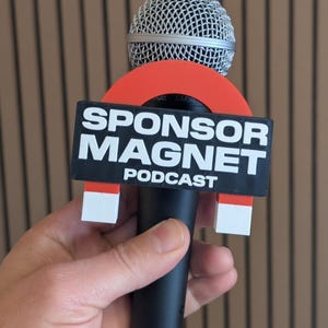 May include: A black microphone with a silver mesh head, featuring a red and black "SPONSOR MAGNET PODCAST" sign. The sign is held in place by white and red magnets.