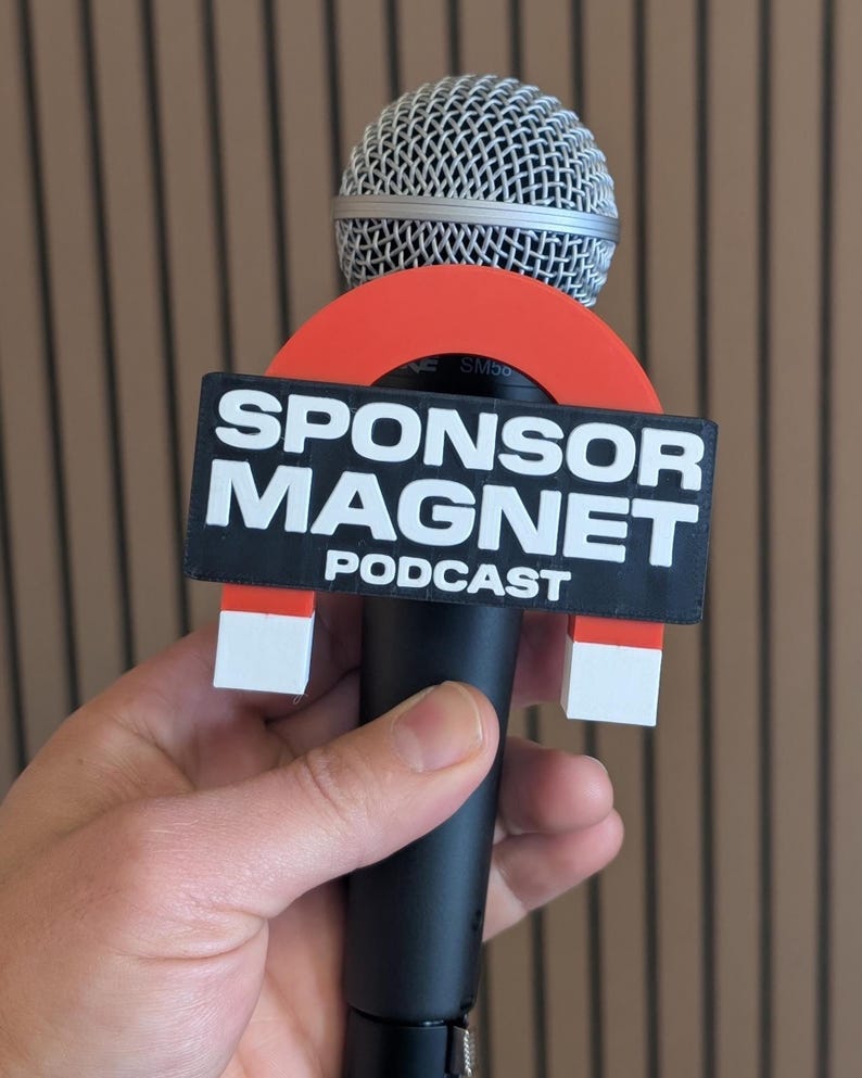 May include: A black microphone with a silver mesh head, featuring a red and black "SPONSOR MAGNET PODCAST" sign. The sign is held in place by white and red magnets.