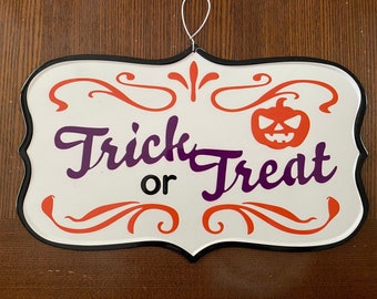May include: White metal sign with black trim and orange and purple lettering. The sign says "Trick or Treat" with a pumpkin face in the upper right corner.