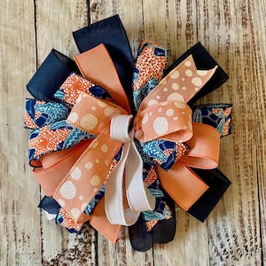 May include: A decorative bow constructed from multiple layers of ribbon. The design features solid navy blue, solid peach, and patterned ribbons with orange, teal, and white abstract shapes, along with a light peach ribbon featuring white polka dots.