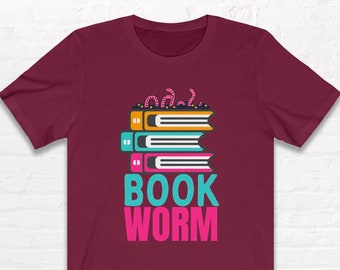May include: A maroon short-sleeve t-shirt featuring a graphic of three stacked books in orange, teal, and pink. Above the books are small pink worms, and below the books is the text "BOOK WORM" in large teal and pink capital letters.