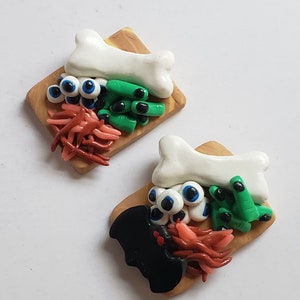 May include: Two miniature wooden chopping boards with Halloween-themed food items. One board has a white bone, green fingers, red worms, and blue eyeballs. The other board has a black bat, green fingers, red worms, and blue eyeballs.