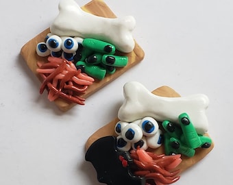 Op de afbeelding: Twee miniatuur houten snijplanken met Halloween-thema voedsel. Een plank heeft een wit bot, groene vingers, rode wormen en blauwe oogballen. De andere plank heeft een zwarte vleermuis, groene vingers, rode wormen en blauwe oogballen.