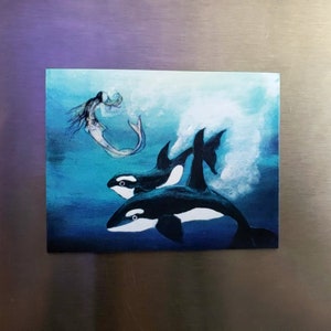May include: A rectangular magnet featuring an underwater scene with two orcas and a mermaid. The orcas are black and white, swimming in blue water. The mermaid has long hair and a fishtail. A white splash is visible.