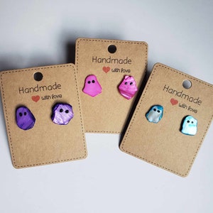 Pode incluir: Três pares de brincos de fantasma feitos à mão em cartão castanho com o texto "Handmade with love". Os brincos são feitos de concha iridescente em roxo, rosa e azul.