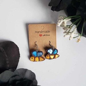 May include: A pair of handmade earrings featuring a mosaic bee design with blue, yellow, and brown accents. The earrings are on a brown card with the text "Handmade with love".