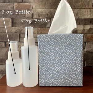May include: Two clear plastic spray bottles with white tops. The smaller bottle is labeled "2 oz. Bottle" and the larger bottle is labeled "4 oz. Bottle". The bottles are sitting on a wooden surface in front of a blue and white tissue box.