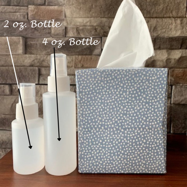 May include: Two clear plastic spray bottles with white tops. The smaller bottle is labeled "2 oz. Bottle" and the larger bottle is labeled "4 oz. Bottle". The bottles are sitting on a wooden surface in front of a blue and white tissue box.