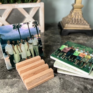 May include: Four square ceramic coasters with photos of people printed on them. The coasters are stacked on top of each other. The top coaster has a photo of a group of people standing in front of a beach at sunset. The coasters are sitting on a wooden stand.