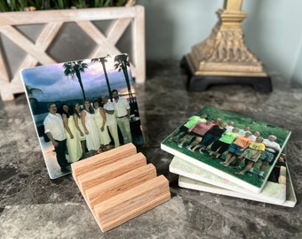 May include: Four square ceramic coasters with photos of people printed on them. The coasters are stacked on top of each other. The top coaster has a photo of a group of people standing in front of a beach at sunset. The coasters are sitting on a wooden stand.