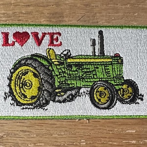 May include: Embroidered patch featuring a green tractor with yellow wheels and the word "LOVE" in red with a heart symbol. The patch has a white background and a green border. The tractor design is detailed.