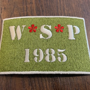 May include: Green embroidered patch with white lettering that reads "W*S*P 1985" with two red embroidered flowers.