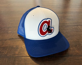 Può includere: Un cappello da baseball bianco e blu con un logo "A" rosso e bianco sul davanti. Il logo presenta una piccola silhouette di una persona seduta su una sedia.