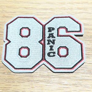 May include: Light blue embroidered patch with the number "86" in a collegiate font. The number has a black and red outline, with the word "PANIC" vertically in the center.