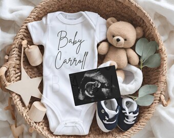 Puede incluir: Un body blanco para bebé con el texto "Baby Carroll" se encuentra en una cesta de mimbre con un osito de peluche, zapatos de bebé y una ecografía. Juguetes de madera y vegetación completan el arreglo, sugiriendo un anuncio de bebé.