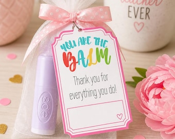 May include: A gift bag with a light purple lip balm, a pink bow, and a tag that reads "YOU ARE THE BALM. Thank you for everything you do!" A white mug with "BEST TEACHER EVER" is in the background.