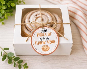 May include: A white gift box with a clear window contains a large cinnamon roll. The box is tied with twine and a round tag that reads "Just rollin' by to say THANK YOU!" in orange and black text. A sprig of green leaves is visible.