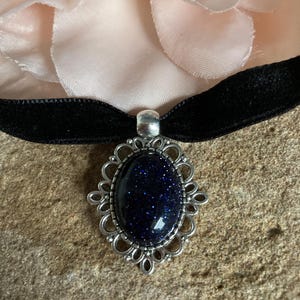 May include: A black velvet choker necklace with a silver-toned pendant. The pendant features a dark blue, oval-shaped stone with sparkling inclusions, set within a decorative, openwork frame. The background includes a light pink flower.