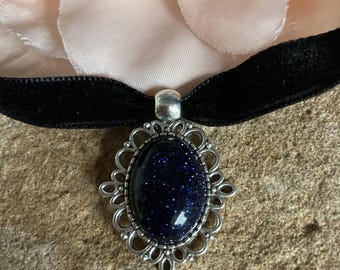 May include: A black velvet choker necklace with a silver pendant. The pendant has a dark blue, oval stone with sparkling inclusions, set in an ornate, openwork frame. A light pink flower is in the background.