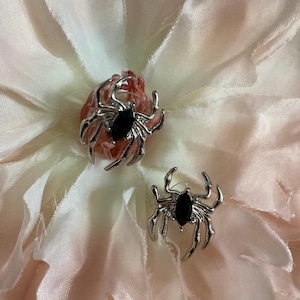 Op de afbeelding: Een paar zilverkleurige spinnen oorbellen met zwarte edelsteen lichamen. De oorbellen zijn geplaatst tegen een zachte, lichtroze en crèmekleurige bloemenachtergrond, wat een opvallend contrast creëert. Een uniek accessoire.