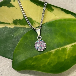 Peut inclure: Un collier en chaîne argent avec un petit pendentif rond. Le pendentif est rempli de paillettes irisées et de petites pièces roses et violettes.