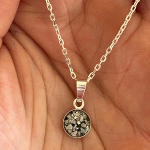 May include: A silver chain necklace with a round pendant. The pendant is filled with small, gray and white pieces of material.