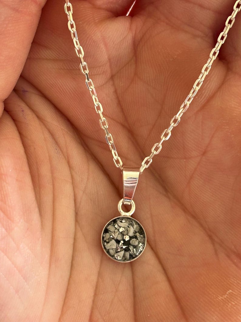 May include: A silver chain necklace with a round pendant. The pendant is filled with small, gray and white pieces of material.