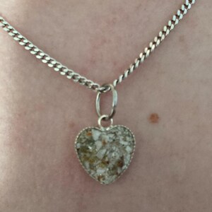 May include: A silver chain necklace with a heart-shaped pendant. The pendant is filled with white and brown sand-like material.