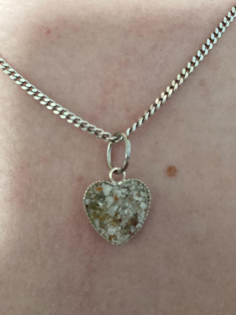 May include: A silver chain necklace with a heart-shaped pendant. The pendant is filled with white and brown sand-like material.