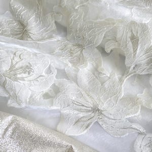 May include: Close-up of white fabric with a floral pattern. The fabric has a delicate, textured appearance with raised floral designs. A section of shimmering silver fabric is visible in the lower portion of the image.
