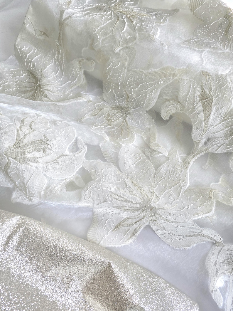 May include: Close-up of white fabric with a floral pattern. The fabric has a delicate, textured appearance with raised floral designs. A section of shimmering silver fabric is visible in the lower portion of the image.