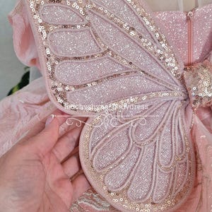 May include: A close-up of a pink fairy wing with gold sequins outlining the edges and veins. The wing is attached to a pink dress with a zipper. The wing is made of a soft, glittery material.
