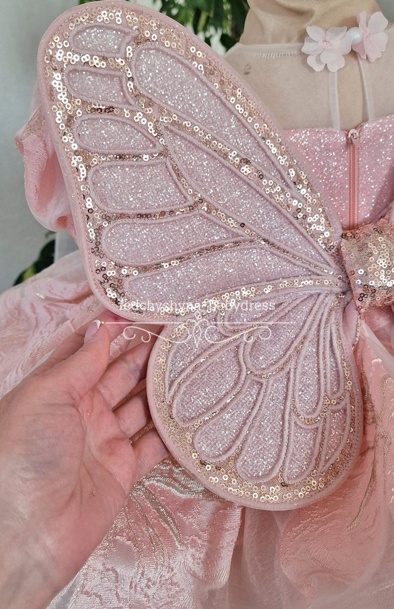 May include: A close-up of a pink fairy wing with gold sequins outlining the edges and veins. The wing is attached to a pink dress with a zipper. The wing is made of a soft, glittery material.