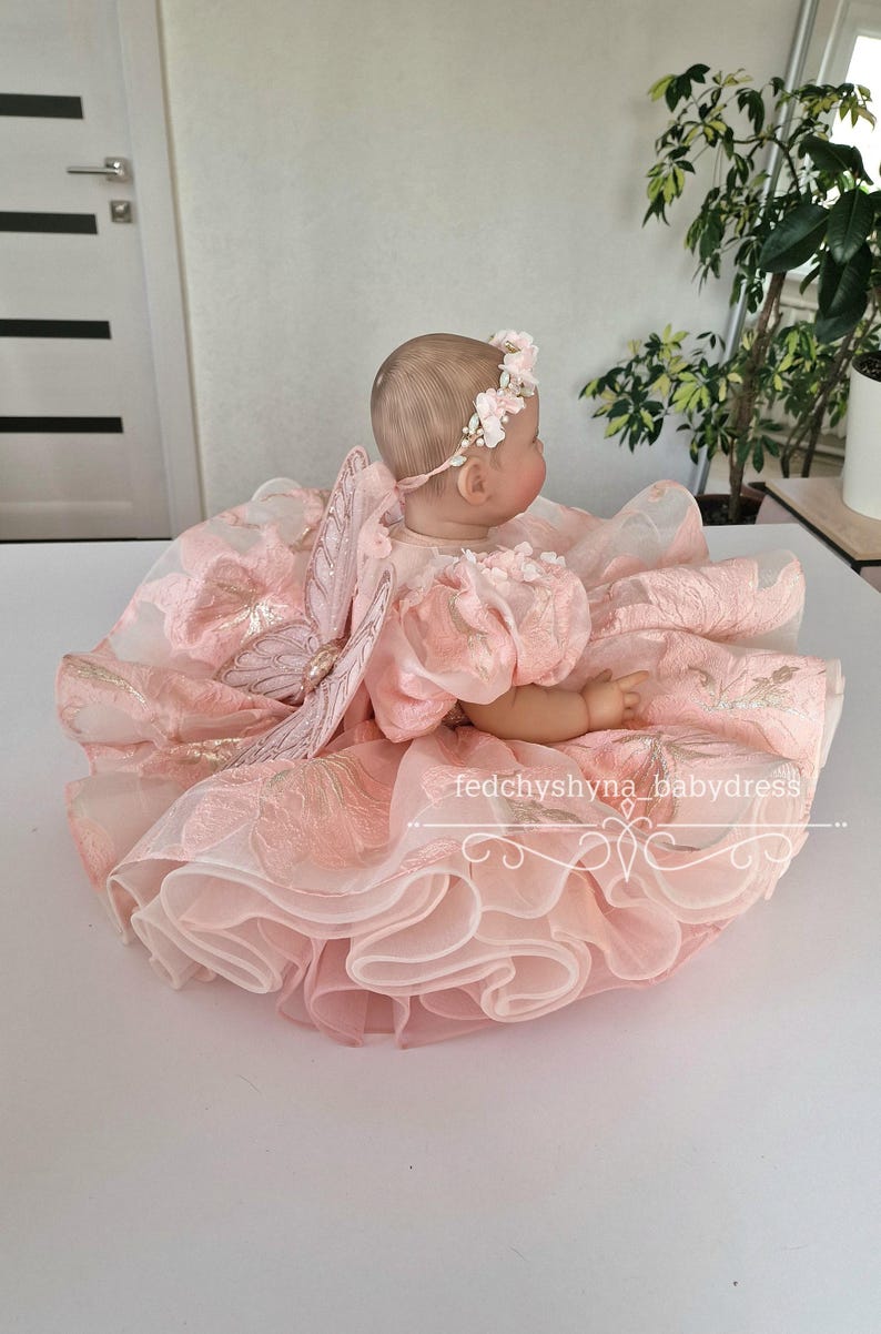 May include: A baby doll wearing a pink dress with a floral headband and butterfly wings. The dress has multiple layers of ruffled fabric and gold accents. The text "fedchyshyna_babydress" is visible.